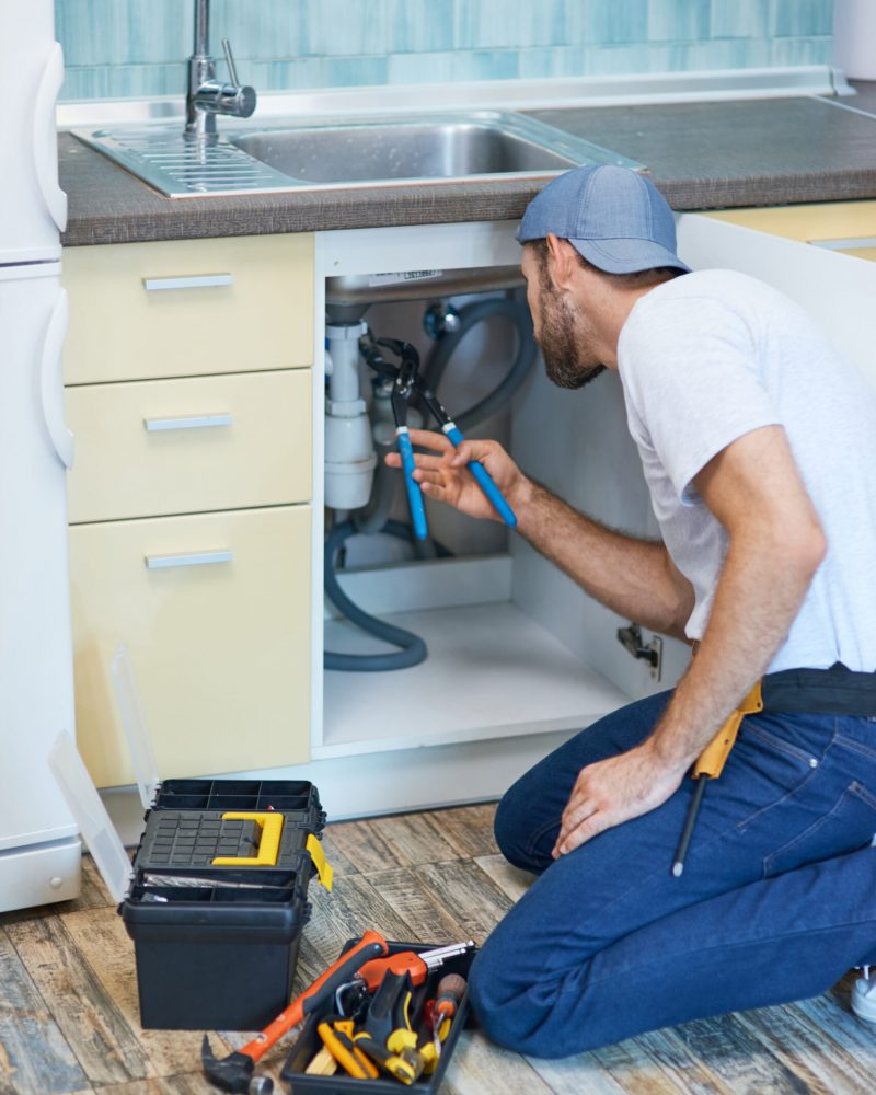 Plumbing troubles. Professional plumber using pipe wrench while examining and fixing sink pipe in the kitchen. Repair service and plumbing concept. Vertical shot
